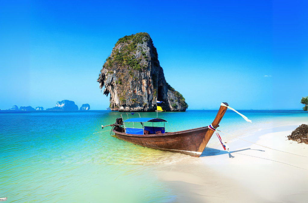 Thailändisches Longtail-Boot an einem weißen Strand vor einem riesigen, bewachsenen Kalksteinfelsen im klaren, türkisen Meer unter blauem Himmel.